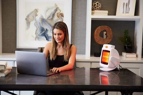 Woman uses the tabletop Infrared Lamp unit at a desk while working on her laptop.
