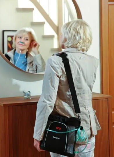 Woman looping a cannula behind her ear while looking into a mirror. She carries a portable oxygen concentrator in a case.