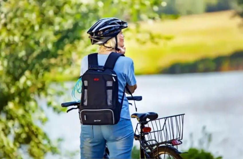 A woman wearing a portable oxygen concentrator in a rucksack standing next to a bike while fastening the straps on her helmet