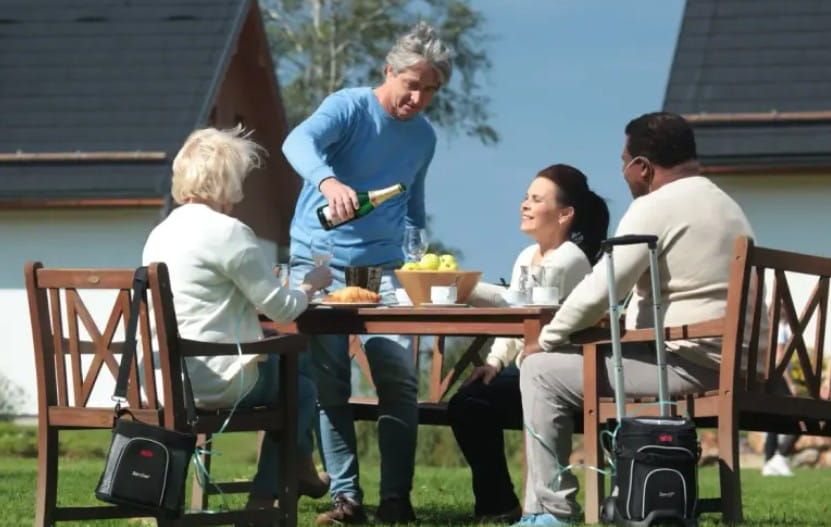 A group of 4 people sits around a table outside. 2 people are using portable oxygen concentrators.