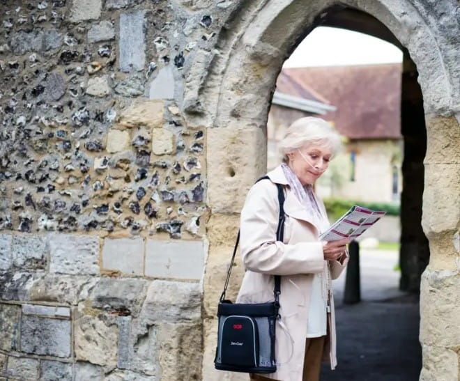 A woman reading a brochure next to an old wall and archway while wearing a portable oxygen concentrator