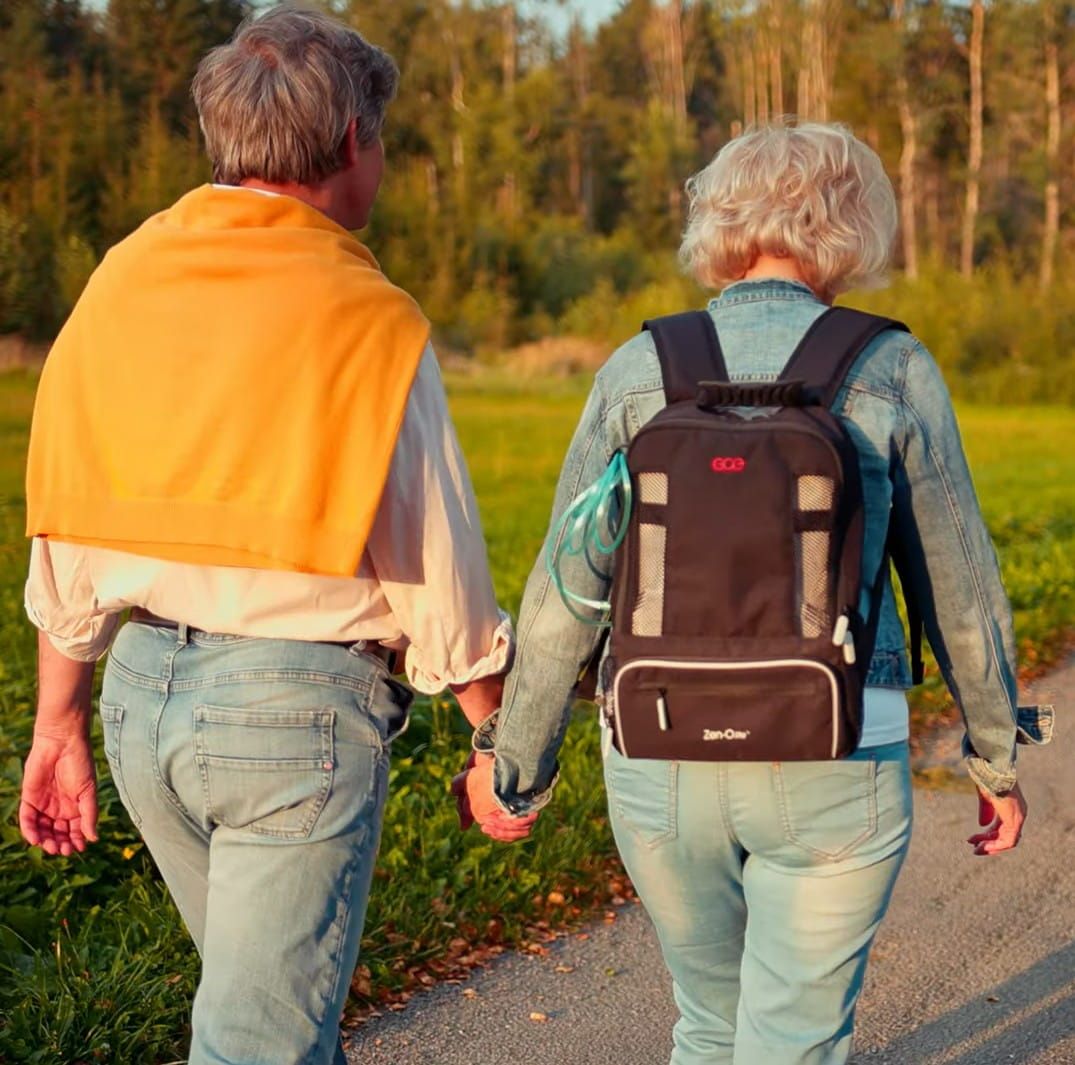 A man and a woman walk hand-in-hand down a road. The woman is wearing a portable oxygen concentrator in a rucksack.