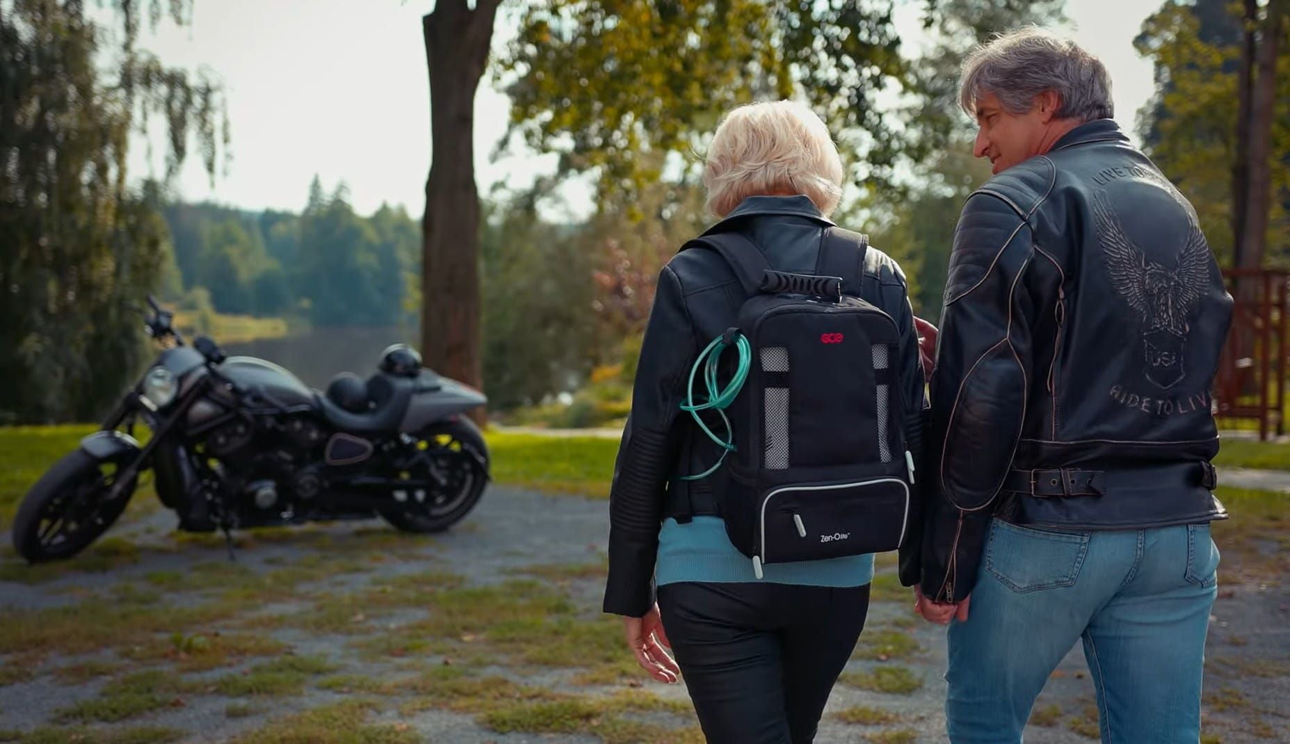 A man and a woman hold hands as they walk toward a motorcycle. The woman is wearing a portable oxygen concentrator in a backpack.