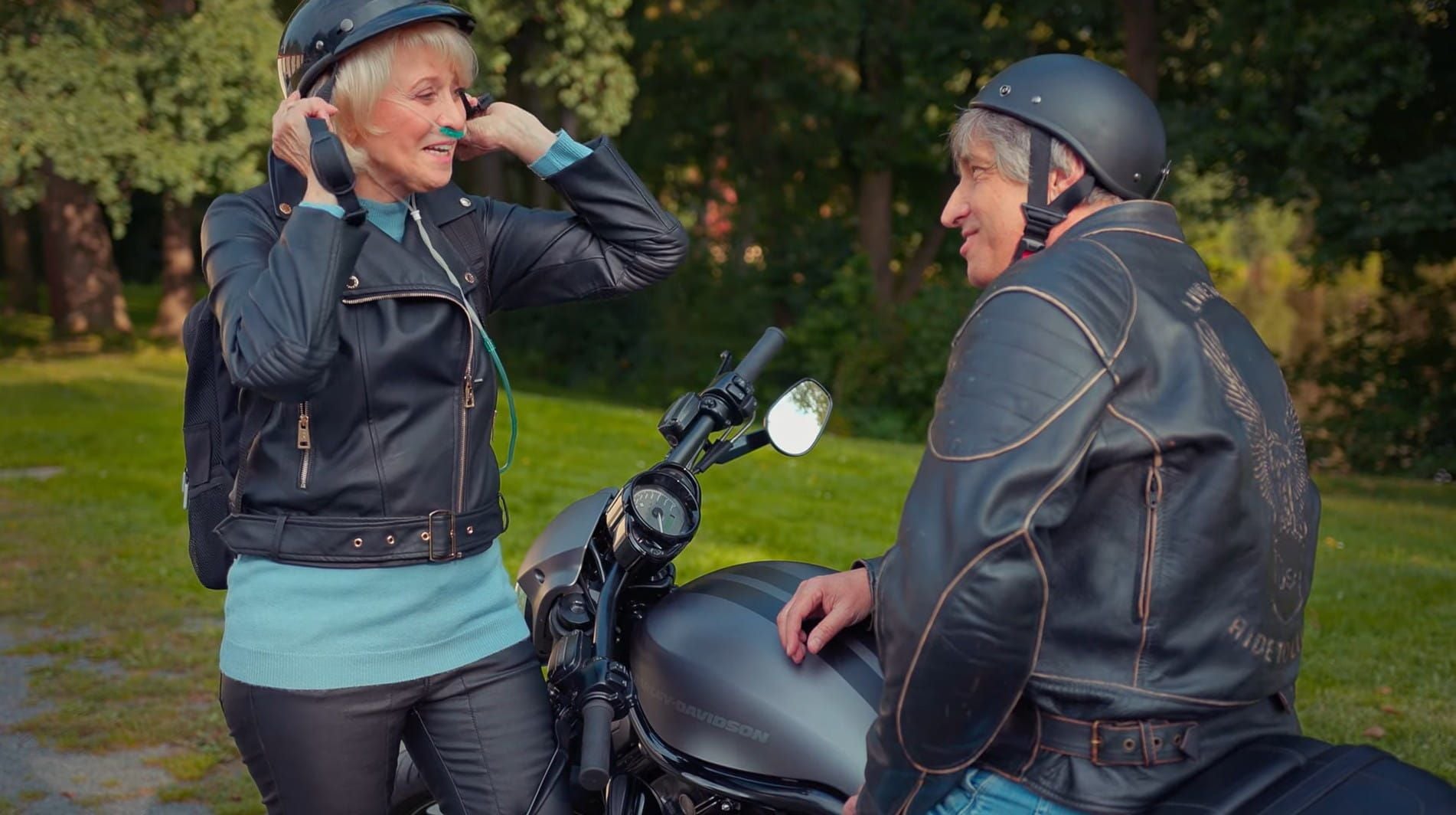 A man is sitting on a motorcycle and talking to a woman who is using a portable oxygen concentrator while putting on a helmet