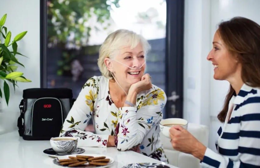 2 Women eating cookies and drinking coffee at a table. One woman using the Zen-O Lite portable oxygen concentrator