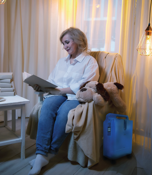 A woman peacefully sits in a chair and reads a book with the concentrator on the floor next to her.